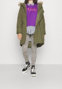 Parka verde oliva con capucha forrada de piel, sobre una sudadera morada con el texto "rose", camisa a rayas, leggings con estampado de cebra y zapatillas negras.