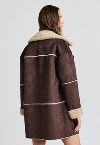 Manteau en suédine marron avec col et poignets en fausse fourrure beige, présentant des coutures horizontales et une coupe ample. Aucun élément métallique ni motif visible.