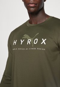 Groene lange mouw shirt van gladde stof, met de tekst "HYROX" en een puma-logo in contrasterende kleuren. Regular fit met een ronde halslijn.