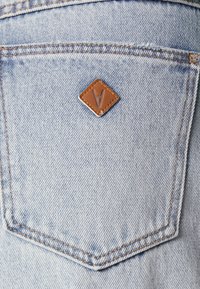 Tecido denim azul claro com um retângulo de couro castanho em forma de losango com um logotipo, posicionado acima de um bolso traseiro com costura em contraste.