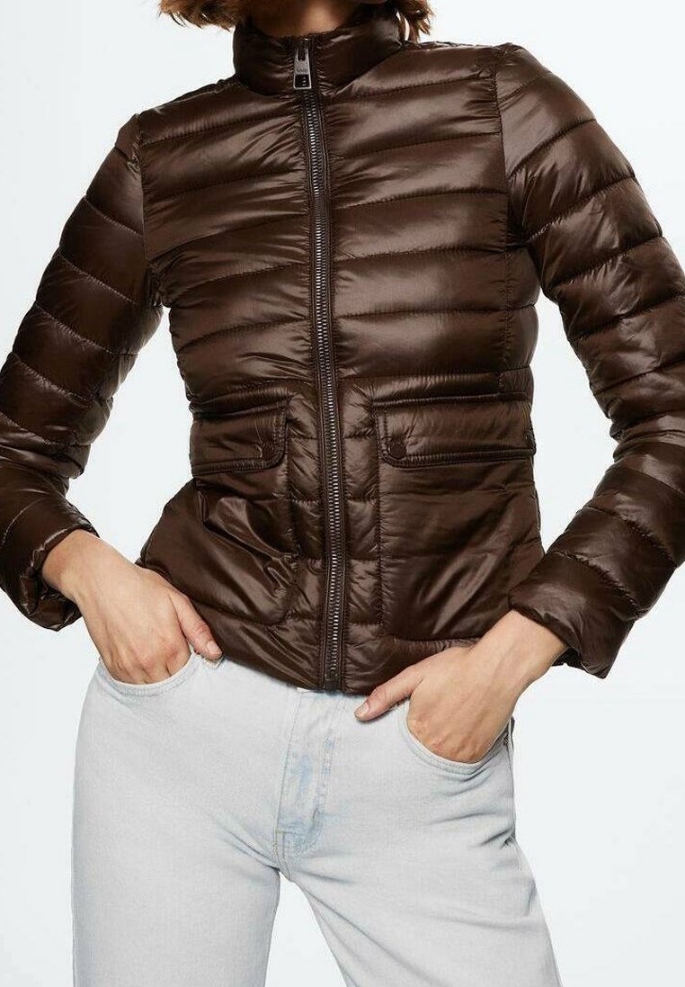 Femme portant une veste matelassée marron foncé ajustée avec des poches avant et un pantalon bleu clair, les mains dans les poches, sur fond blanc.