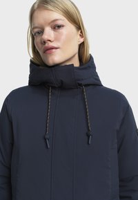 Marineblauwe pufferjas met een hoge kraag, gouden trekkoorden en zijzakken. Heeft een gladde, matte textuur en subtiele stikdetails.