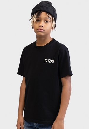 Unge person iført en sort hue og sort t-shirt med hvide japanske tegn på en ensfarvet lys baggrund.