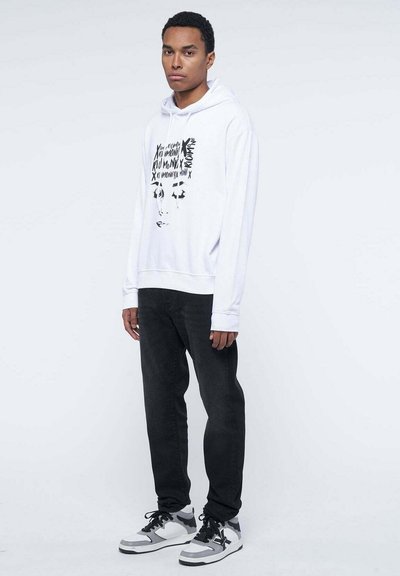 Felpa bianca con stampa grafica nera, maniche lunghe e tasca a marsupio frontale, abbinata a pantaloni neri e sneakers grigie e bianche.