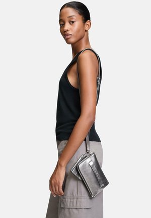 Metallisch silberne Umhängetasche mit glatter Textur, ausgestattet mit einem Reißverschluss und einem abnehmbaren Trageriemen, getragen von einem Model in einem schwarzen Tanktop.