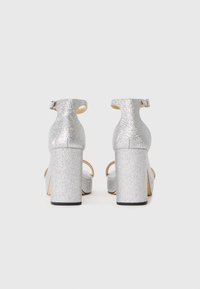 Silberglänzende, glitzernde Sandaletten mit hohen Absätzen, Knöchelriemen und Blockabsätzen, von hinten auf weißem Hintergrund gezeigt.