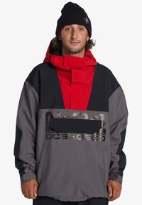 Šedý, červený a čierny pulóver s vysokým golierom, polovičným zipsom vpredu a veľkou prednou kapsou. Obsahuje potlačené logo a reflexné detaily.