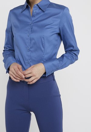 Femme portant une chemise bleue cintrée à boutons avec manches longues et un pantalon bleu assorti, mains légèrement jointes au niveau de la taille.