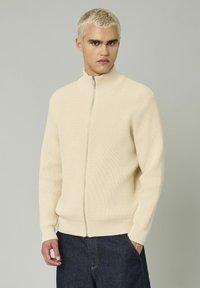 Gilet zippé côtelé beige avec un col montant, des poignets ajustés et une ceinture. Texture lisse et couleur neutre, porté par-dessus un jean en denim foncé.