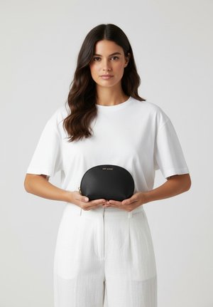 Zwarte gelapte leren pouch met een afgeronde vorm, gouden rits en een geperst logo. Vastgehouden door een persoon die een wit T-shirt en een broek draagt.