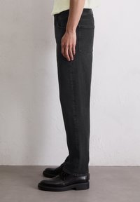 Osoba ubrana w czarne dżinsy o prostym kroju i czarne skórzane buty formalne stoi przy gładkiej białej ścianie.