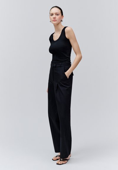 Femme debout sur un fond uni, portant un top noir sans manches, un pantalon noir taille haute à jambes larges, et des sandales noires style tongs, une main dans la poche.