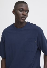 Tricou cu mânecă scurtă de culoare albastră închisă, confecționat din bumbac, cu un buzunar pe piept și guler rotund, având o textură netedă.