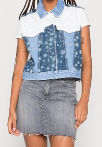 Jeansowa kamizelka z białym kołnierzem, przodem w kwiatowy wzór oraz dwoma kieszeniami na piersiach; w zestawie z postrzępioną szarą jeansową spódnicą.