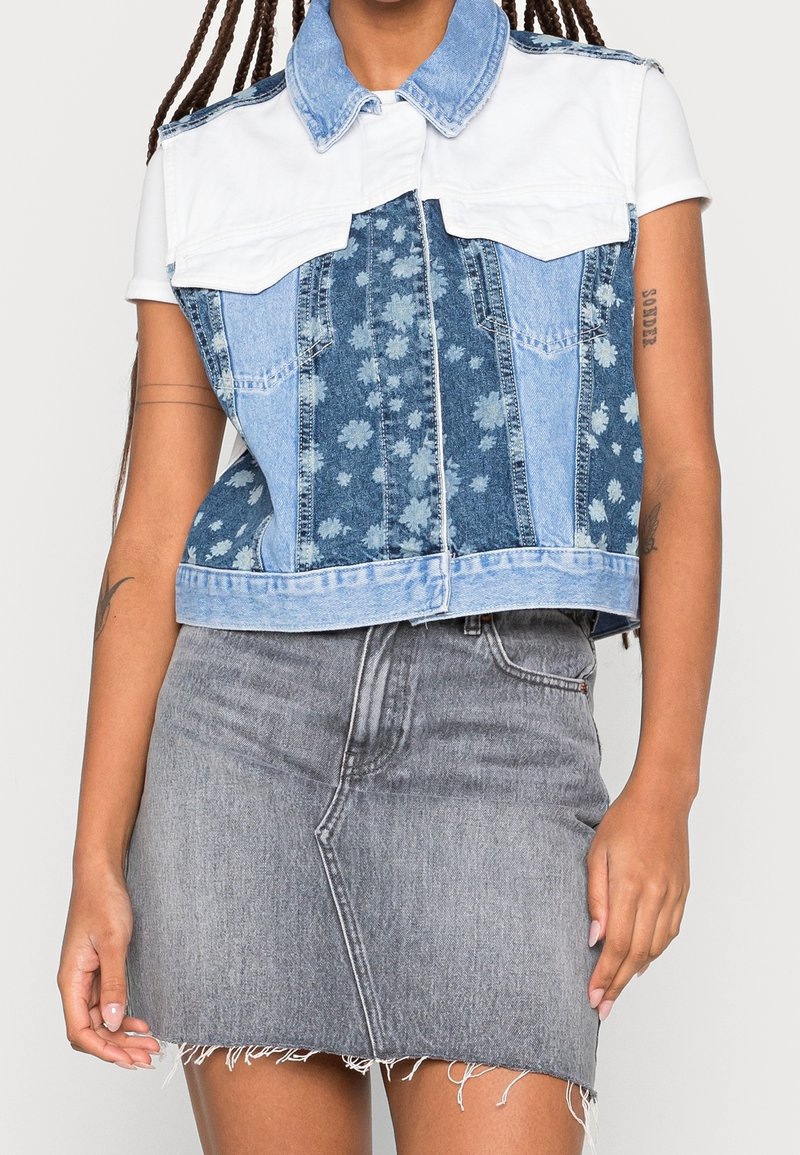 Jeansowa kamizelka z białym kołnierzem, przodem w kwiatowy wzór oraz dwoma kieszeniami na piersiach; w zestawie z postrzępioną szarą jeansową spódnicą.