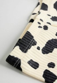 Tejido acanalado en beige con patrón de manchas de vaca en negro; presenta costuras visibles y superficie texturizada; se muestra la esquina de una prenda.