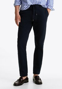 Chinos blu navy con una vestibilità rilassata, dotati di vita regolabile con cordino, tasche anteriori e texture liscia, abbinati a mocassini marroni.