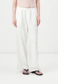 White, lightweight linen pants with a relaxed fit, elastic waistband, and side pockets, worn with black flip-flops on a neutral background.