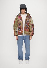 Puffy, multicolored jacket with abstract patterns, featuring a zip front and side pockets, paired with light blue jeans and white sneakers.