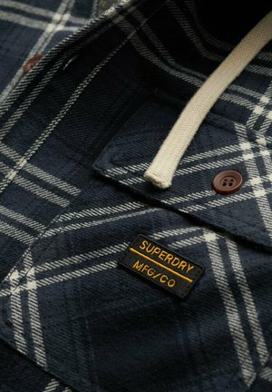 Camisa de franela azul marino con un patrón de cuadros en blanco y gris, que presenta un bolsillo con botones y una etiqueta negra con texto amarillo. Textura suave.