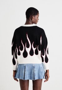 Μαύρο πουλόβερ με σχέδιο φωτιάς σε κόκκινο και λευκό, σε oversized γραμμή, με ριμπ μανσέτες και στρογγυλή λαιμόκοψη. Συνδυάζεται με μια ελαφριά τζην φούστα με πτυχώσεις.