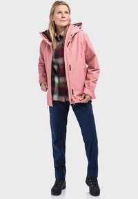 Giacca impermeabile rosa con cappuccio, indossata sopra una camicia a quadri verde e rossa, abbinata a pantaloni blu e scarponi da trekking neri.