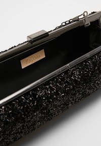 Pochette noire avec un extérieur texturé composé de petites perles. L'intérieur est doublé d'un tissu sombre, avec une fermeture métallique et un détail de logo.