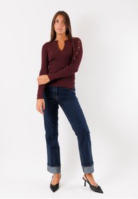 Maglietta lunga a maniche con scrittura in borgogna, scollo a V e dettagli con bottoni sulla manica sinistra, abbinata a jeans blu scuro con risvolto e tacchi neri.