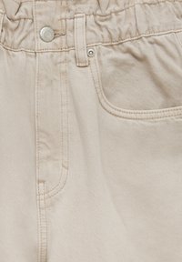Gros plan sur un tissu denim beige montrant une ceinture élastique, un bouton métallique, une passants de ceinture et une poche avant avec des détails de couture.