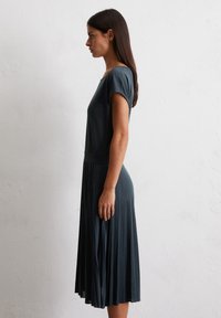 Abito plissettato di un verde scuro con maniche corte, scollatura tonda e corpetto aderente. Tessuto morbido e liscio con una silhouette fluida.