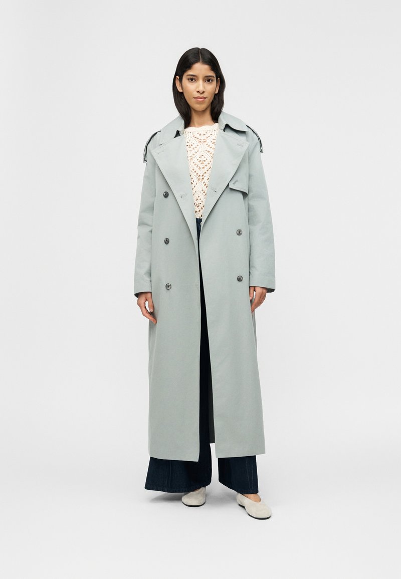 Γυναίκα που φοράει ελαφρύ γκρι μακρύ διπλομπουκ λωτό παλτό, λευκή μπλούζα με κροσέ, σκούρα φαρδιά παντελόνια και λευκά μπαλαρίνες, στέκεται σε λευκό φόντο.