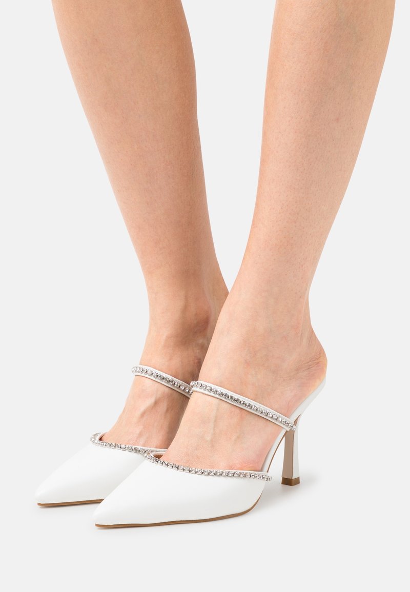 Mules blanches à talons hauts à bout pointu avec deux fines brides ornées de strass, portées pieds nus sur un fond uni.