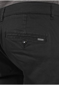 Zbliżenie tylnej kieszeni czarnych spodni z zapięciem na guzik i małą, paskowaną metką materiałową nad szczeliną kieszeni.