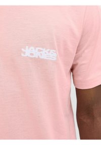 Lyserød bomulds t-shirt med korte ærmer, der har et hvidt logo trykt på venstre side. Blød tekstur og afslappet design.