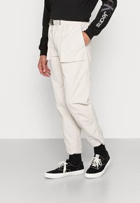 Pantaloni cargo beige chiaro con cordino e cintura, tasche laterali e una texture sintetica liscia. Abbinati a sneakers nere.