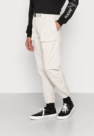 Pantaloni cargo beige chiaro con cordino e cintura, tasche laterali e una texture sintetica liscia. Abbinati a sneakers nere.