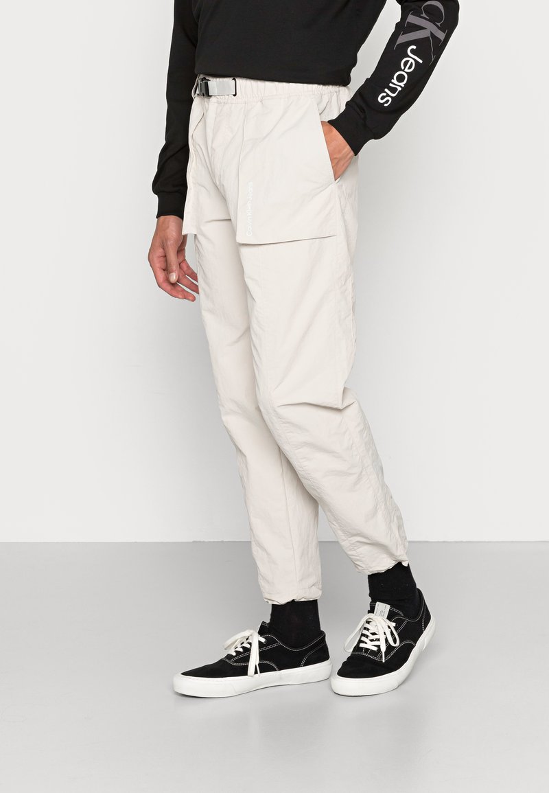 Pantaloni cargo beige chiaro con cordino e cintura, tasche laterali e una texture sintetica liscia. Abbinati a sneakers nere.