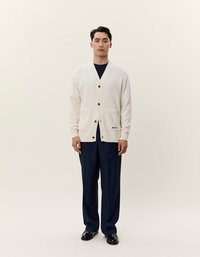 Cardigan crema con maglia texture, caratterizzato da un collo a V, chiusura con bottoni, due tasche frontali e un dettaglio del marchio. Indossato sopra pantaloni blu navy.