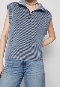 Ärmelloses blau-weiß gestreiftes Oberteil mit einem Reißverschlusskragen, gerippter Textur, lockerer Passform, getragen zu hellblauen High-Waist-Jeans.