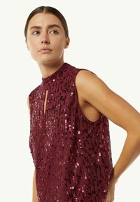 Burgunderfarbenes ärmelloses Oberteil mit hohem Kragen, das mit einem Schlüssellochdetail und vertikalen Paillettenmustern in unterschiedlichen Farbtönen verziert ist.