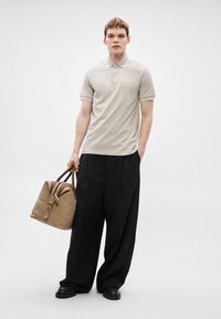 Μπεζ πόλο μπλουζάκι με κοντά μανίκια, συνδυασμένο με φαρδιά μαύρη παντελόνα. Το μοντέλο κρατά μια ταμπά τσάντα με δερμάτινες λαβές και λεπτομέρεια με πόρπη.