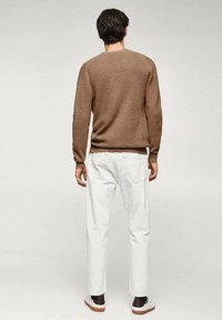 Hnědý pletený svetr s kulatým výstřihem, nošený přes bílé kalhoty. Outfit má volný design s viditelnou texturou a detaily střihu.