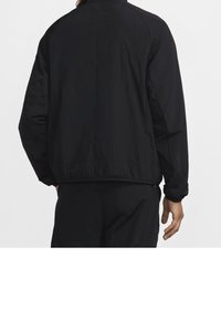 Persoon in een zwarte lange-mouwen jack en bijpassende broek, van achteren weergegeven tegen een effen witte achtergrond.