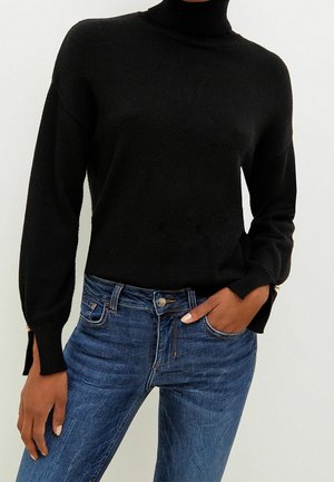Personne portant un pull noir à col roulé manches longues et un jean bleu, une main dans la poche du jean et l'autre reposant le long du corps.