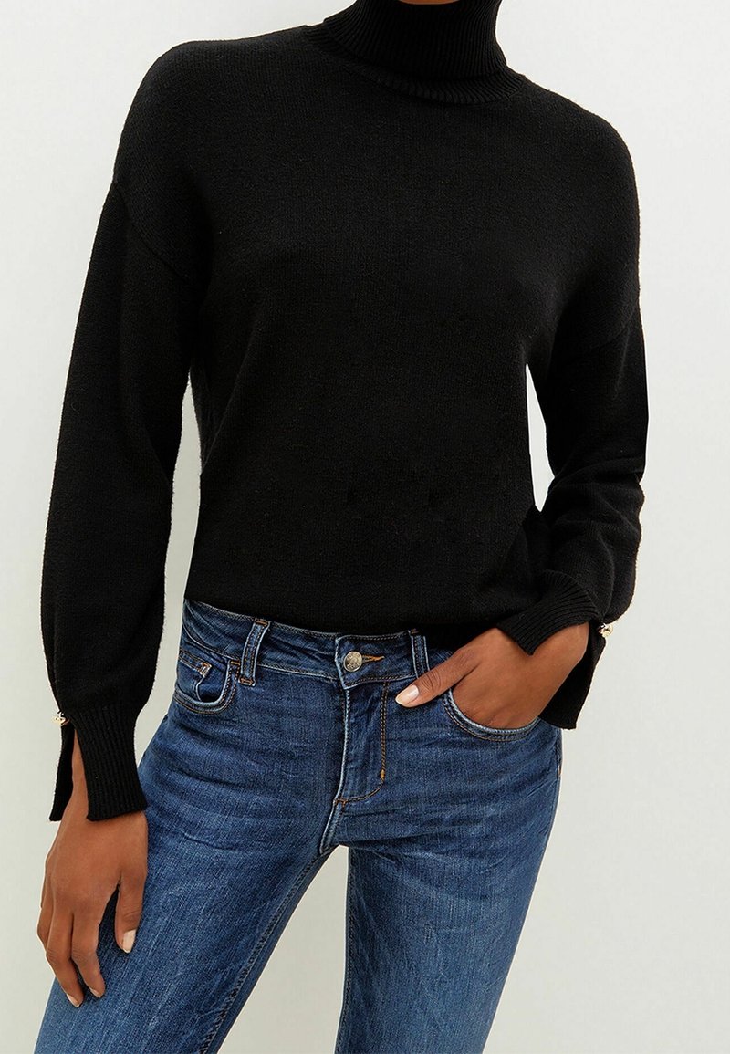 Personne portant un pull noir à col roulé manches longues et un jean bleu, une main dans la poche du jean et l'autre reposant le long du corps.