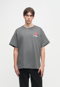 Pelēka īsās piedurkņu T-kreklā izgatavots no kokvilnas, ar sarkanu logo un zivs grafiku kreisajā pusē, brīvā, kvadrātveida piegriezumā.