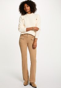 Maglione a coste crema con dettagli dorati, abbinato a pantaloni a zampa d'elefante color marrone chiaro con tasche frontali e chiusura con bottoni, indossato con stivali alla caviglia beige.