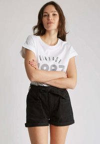 T-shirt blanche avec imprimé "VINTAGE 1987", associée à un short en denim noir taille haute avec poches avant et ceinture nouée.