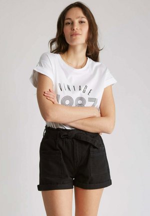 T-shirt blanche avec imprimé "VINTAGE 1987", associée à un short en denim noir taille haute avec poches avant et ceinture nouée.
