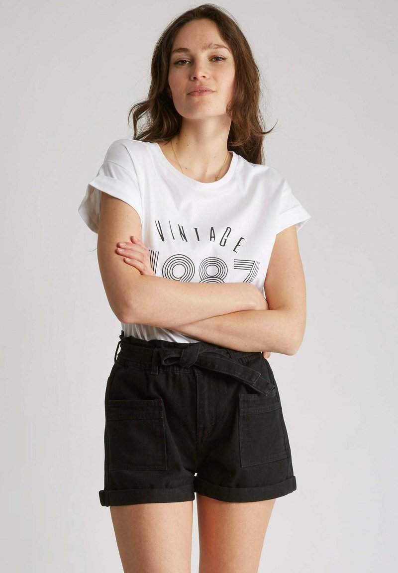 T-shirt blanche avec imprimé "VINTAGE 1987", associée à un short en denim noir taille haute avec poches avant et ceinture nouée.